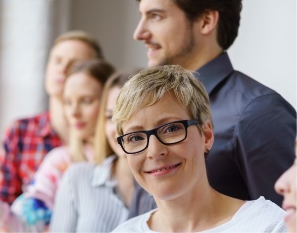 Im Hintergund eine Gruppe junger Menschen und eine Frau schaut lchelnd direkt in die Kamera